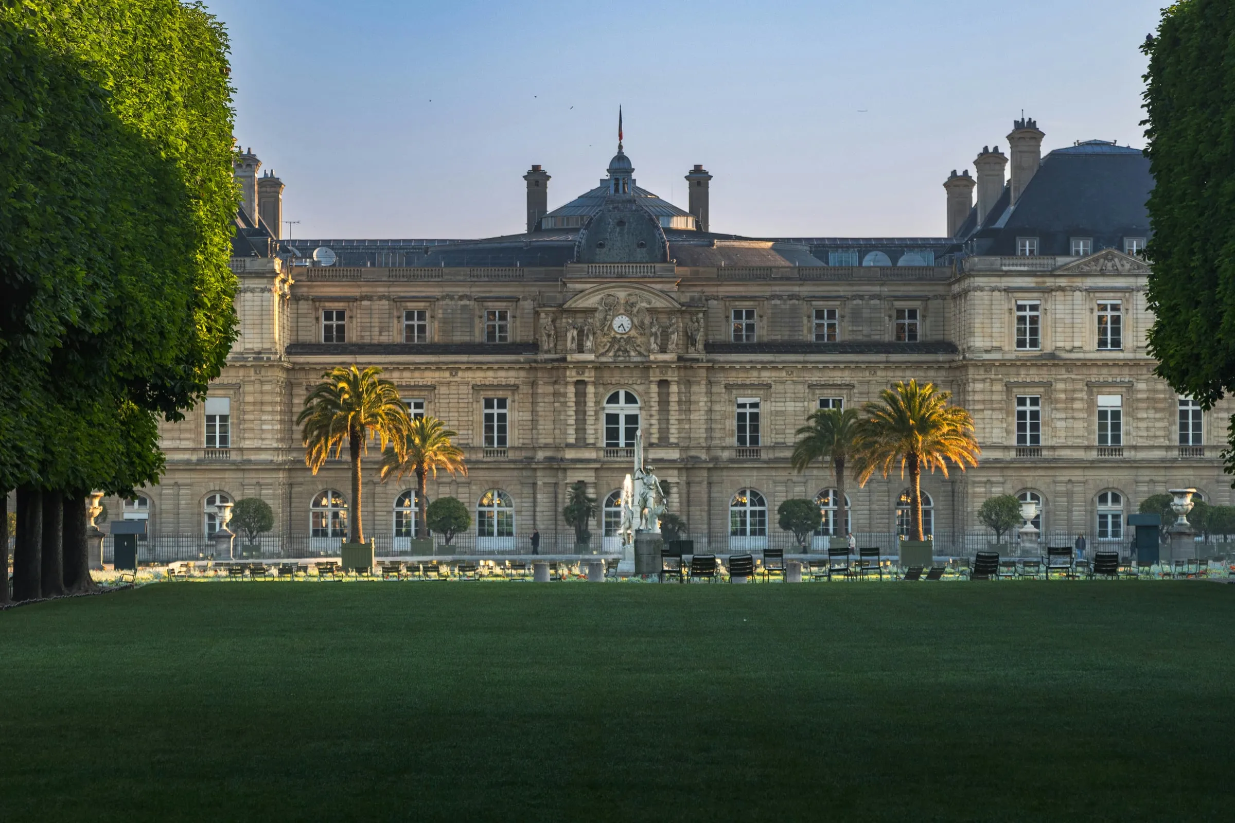 The garden front of the Luxembourg Palace in early light across a wide lawn, palm trees in tubs and a row of green chairs along the terrace, clipped trees framing the view on either side