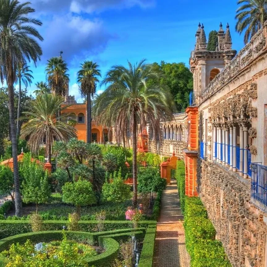 The Real Alcázar's gardens: box-hedged beds and a brick path beside a rusticated stone gallery with ornate turrets, tall palms and an orange-washed pavilion beyond under a blue sky