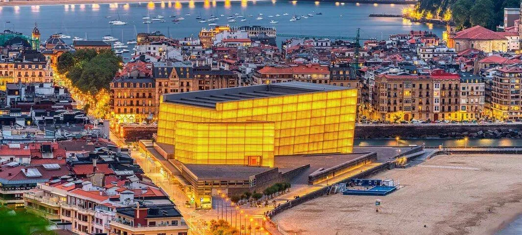 The Kursaal's two glass cubes glowing amber at dusk at the mouth of the river, a beach below them and the lit streets and rooftops of the city behind, with boats moored in the bay beyond