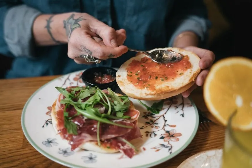 A tattooed hand spreading grated tomato on a toasted roll, beside another topped with jamón and rocket on a flowered plate, with a cut orange in front