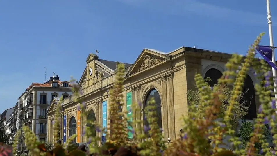 The market's sandstone façade with a clock in its pediment and tall arched windows hung with banners, seen through flowering plants under a blue sky
