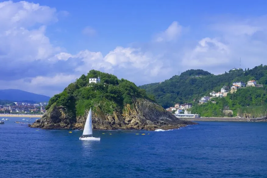 A white sailing yacht off Santa Clara island, a wooded rock with a small white building on top, with the wooded slopes and houses of Monte Igeldo behind and a long beach at the far left