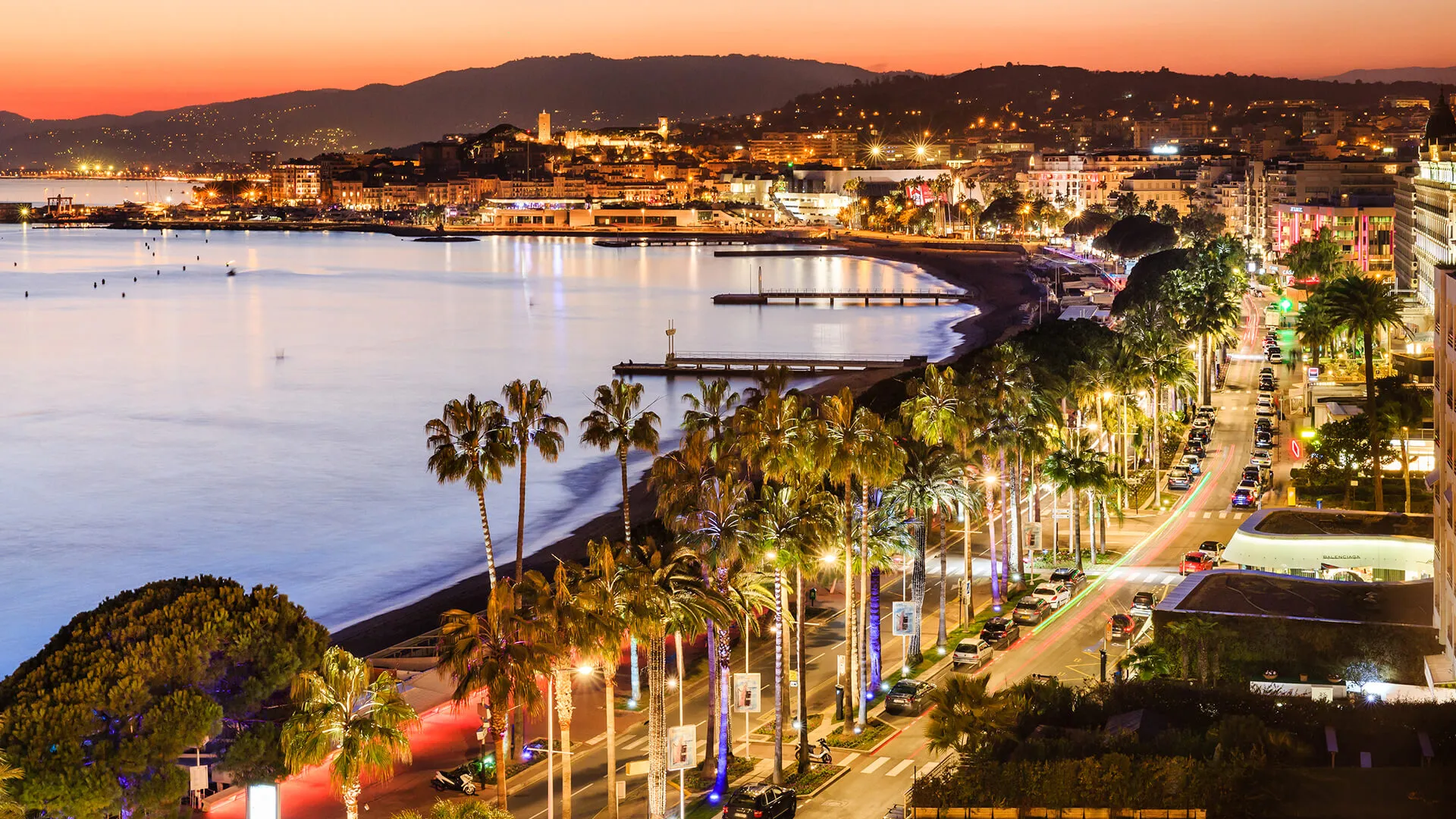 The Croisette in Cannes at dusk: a curve of beach and jetties along a still bay, palms lit along the boulevard and its lanes of traffic, hotels and shops on the right, and the old town on its hill with its tower in the distance, under a pink sky