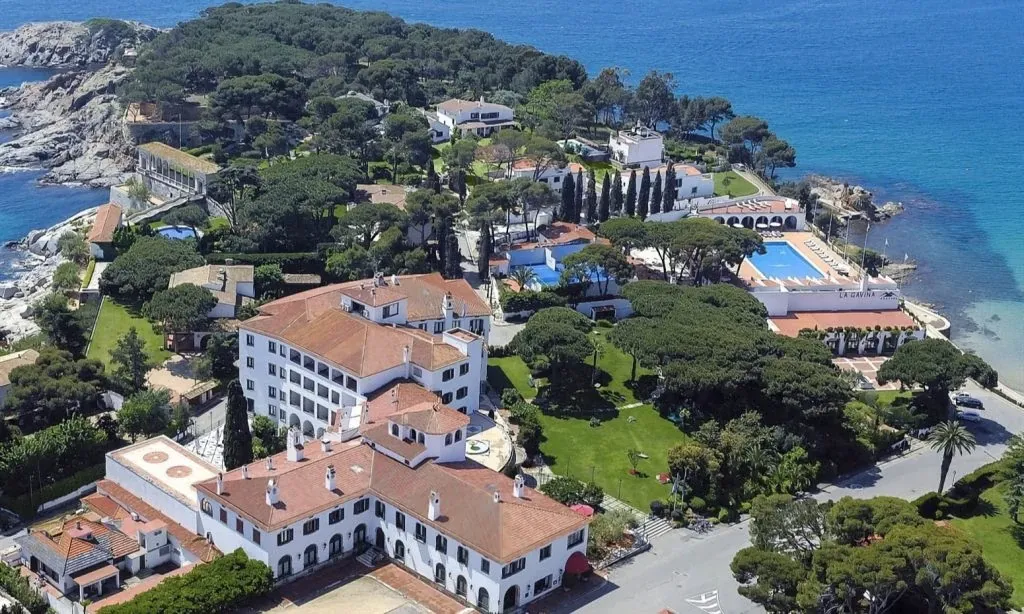 Hostal de La Gavina from the air, white with terracotta roofs among pines above the sea