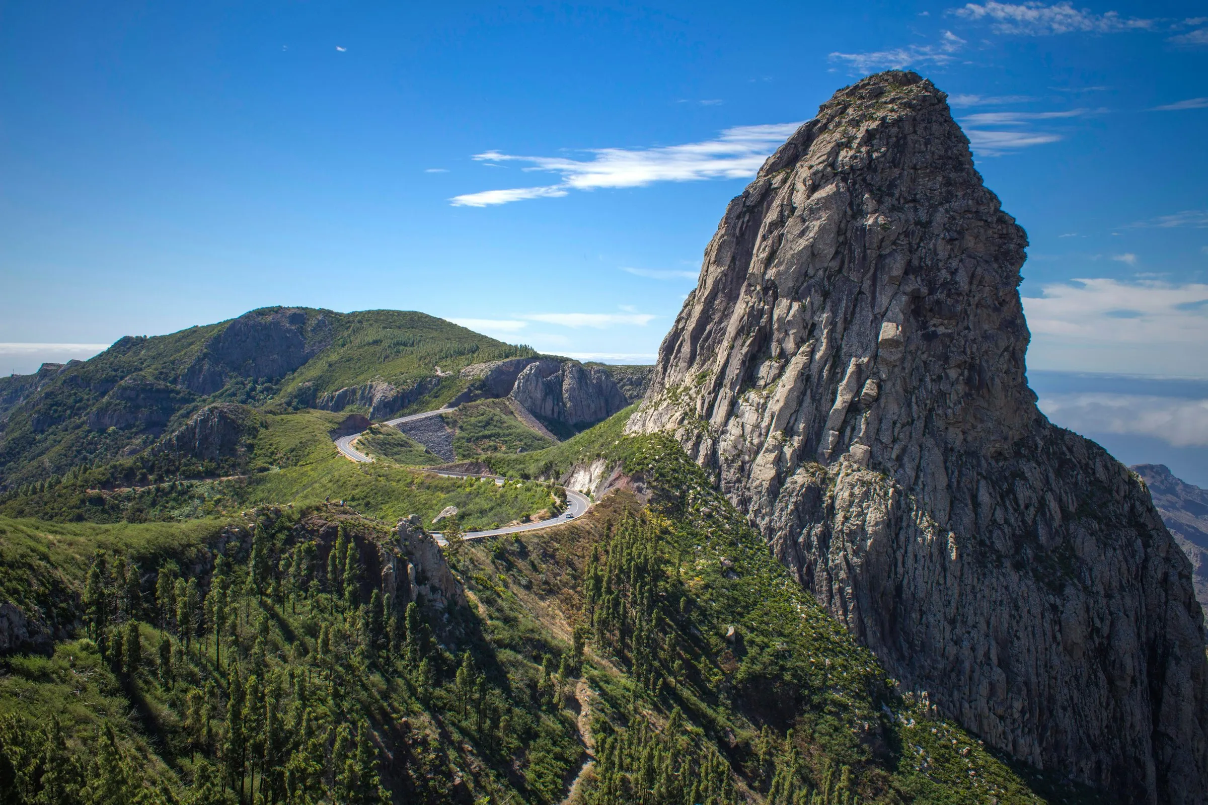 The sheer grey tower of Roque de Agando rising above pine-covered slopes in the heart of La Gomera, a mountain road winding past it under a deep blue sky