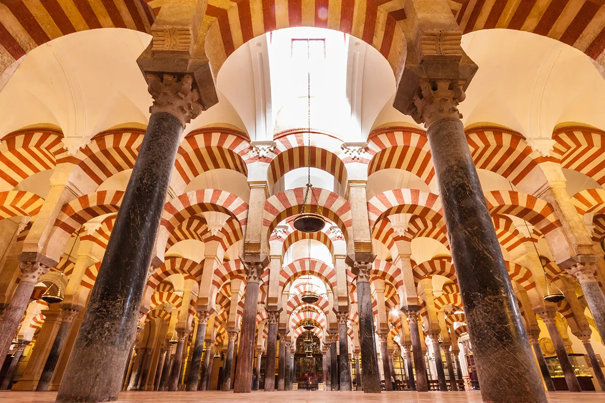 Looking up between dark marble columns into the Mezquita-Catedral's prayer hall, rows of double horseshoe arches striped in red brick and pale stone running into the distance