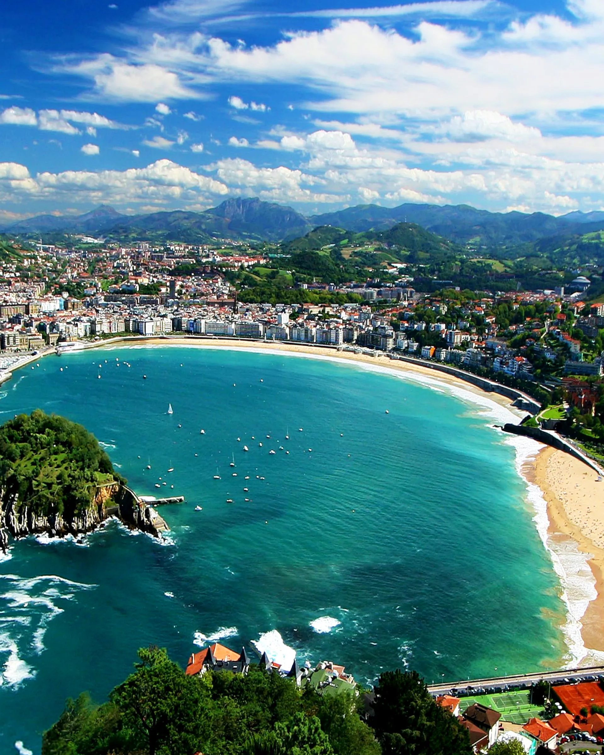 La Concha bay from high above: Santa Clara island and its jetty on the left, sailing boats at anchor in turquoise water, the long curve of sand and the city's seafront behind, green hills and mountains beyond under white clouds