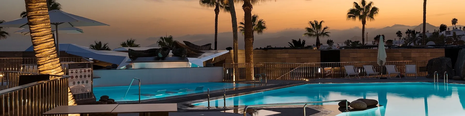 The pool at La Isla y el Mar at sunset, lit from below, with palms, folded white canopies and the volcanic hills beyond