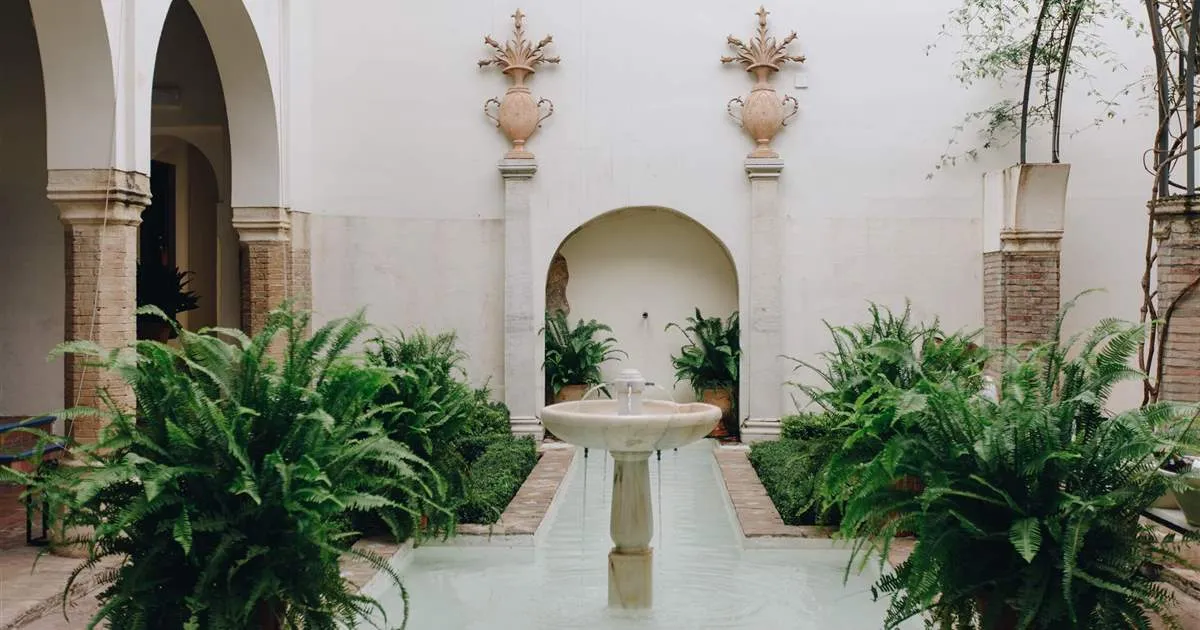 A patio at Las Casas de la Judería: a white marble fountain playing into a long, narrow pool between beds of ferns, brick arches on stone columns to the left, and a whitewashed wall behind with an arched niche between two pilasters topped by stone urns