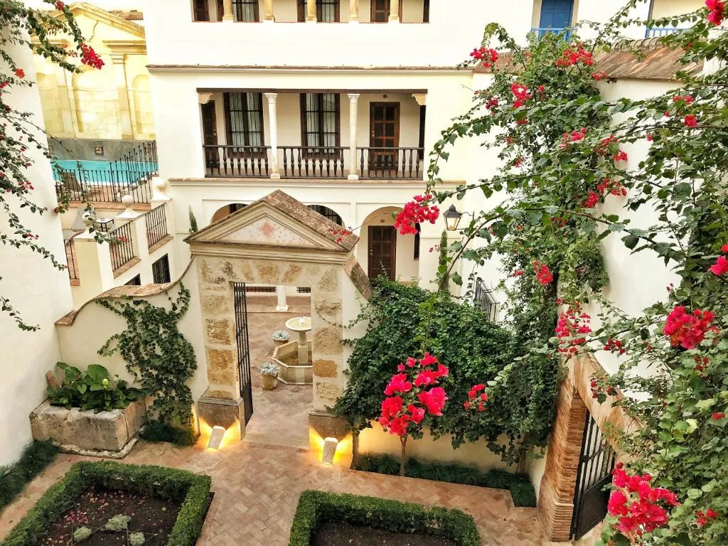 A patio seen from above: a carved stone doorway lit at its foot, beds of low hedges, pots of red geraniums and a bougainvillea in flower, white walls with iron balconies around it