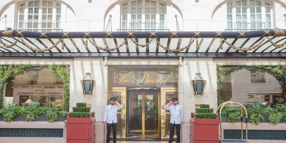 The entrance of Le Bristol Paris under a canopy trimmed with gold rope, two doormen at the glass doors, brass lanterns on the stone and clipped box in red planters