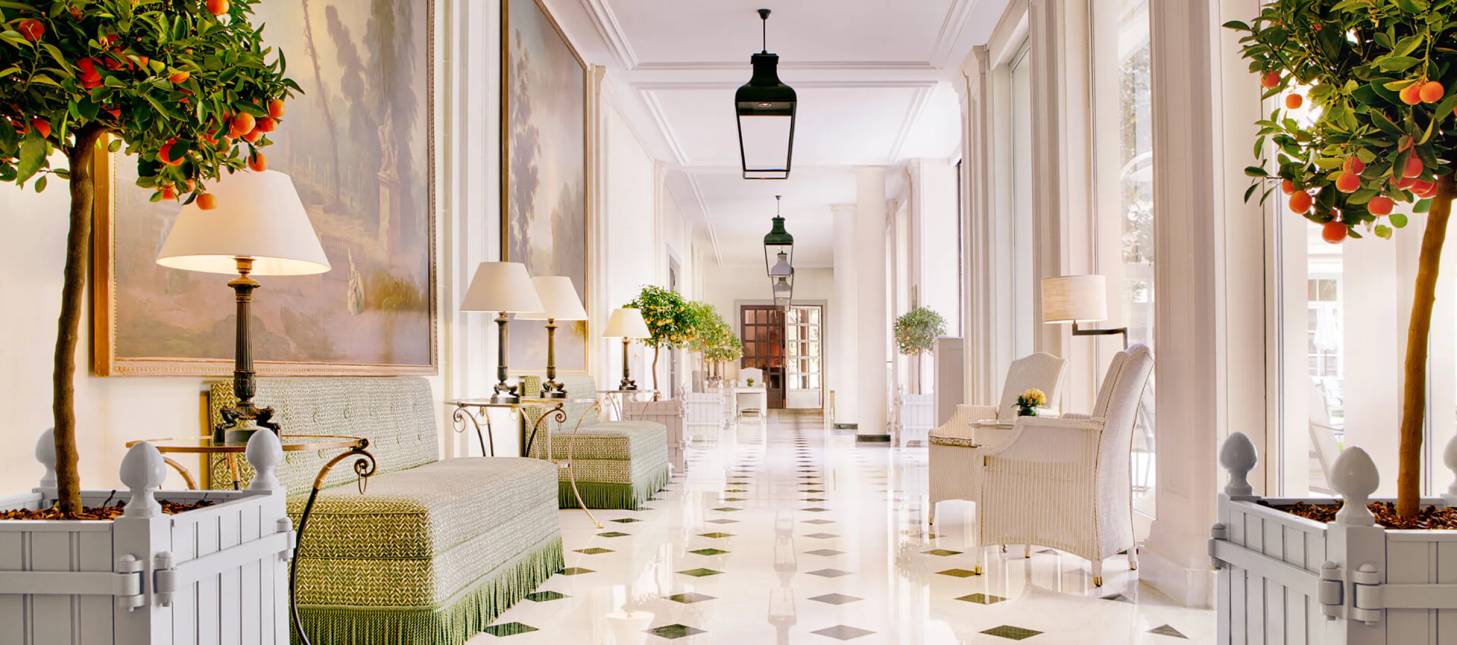 A long white gallery with a marble floor inlaid with green diamonds, green banquettes and bronze lamps beneath large landscape paintings on one side, white wicker armchairs by tall windows on the other, lanterns hanging overhead and orange trees in white planters in the foreground
