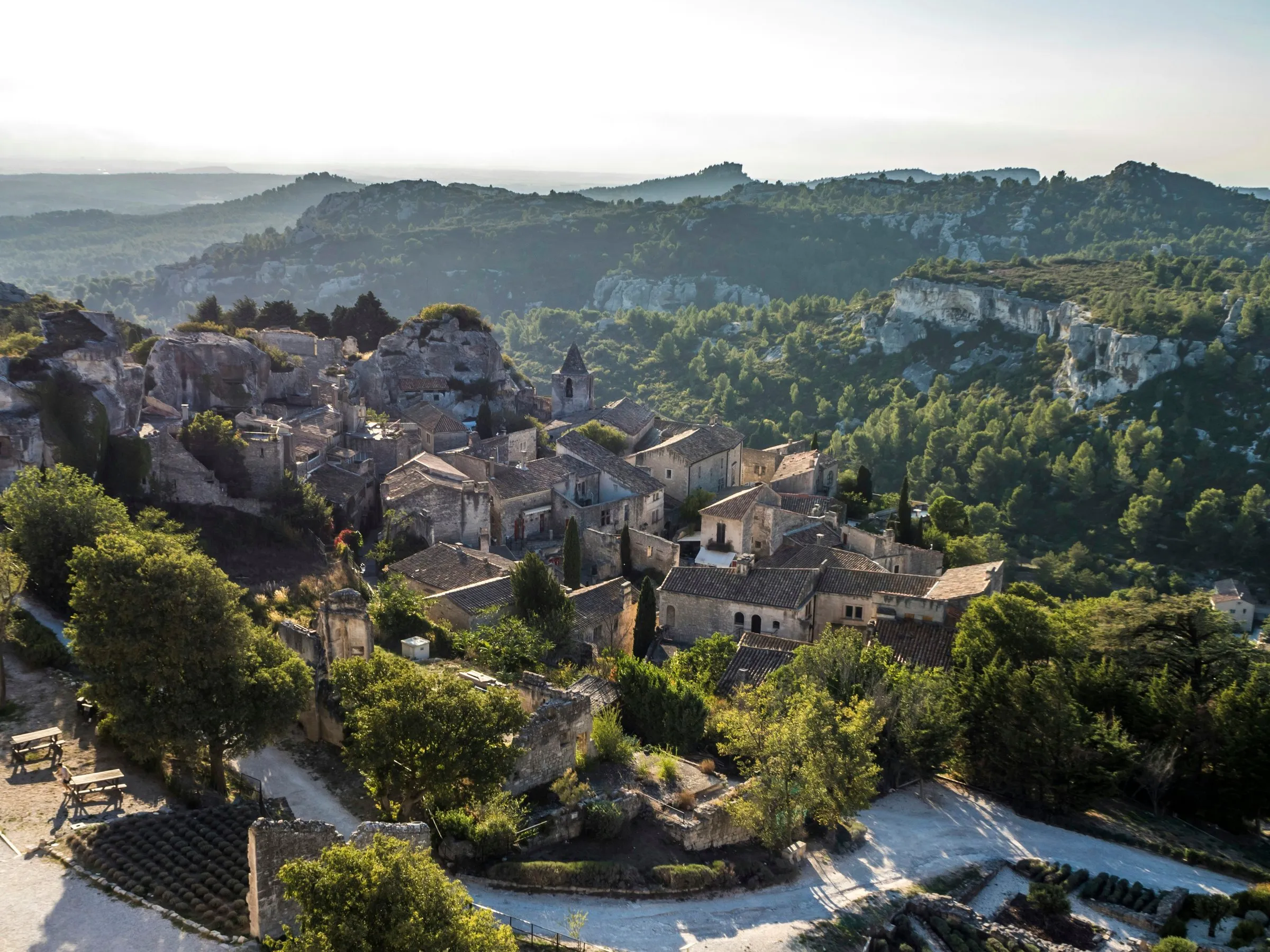 The stone roofs of Les Baux-de-Provence in late golden light, seen from the castle plateau, the church tower among them and the pine-covered rocks of the Val d'Enfer behind
