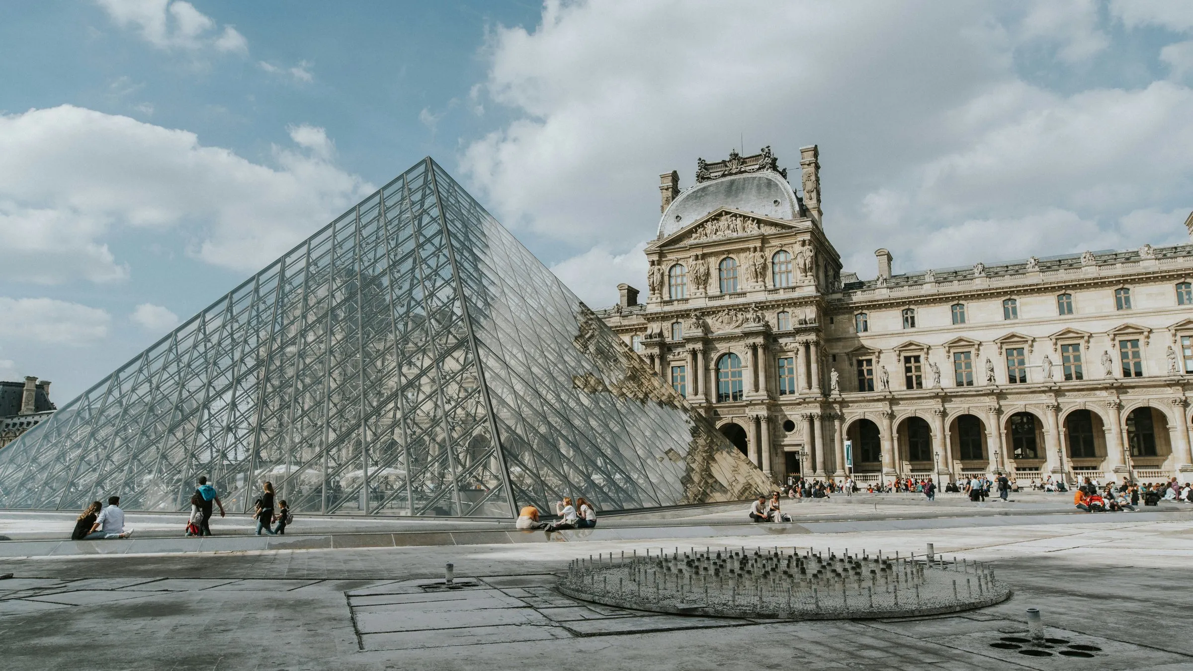 The Louvre's glass pyramid in the Cour Napoléon under a cloudy blue sky, the palace's domed pavilion and long arcaded wing behind it to the right, a dry fountain basin on the paving in the foreground and a few visitors small in the distance