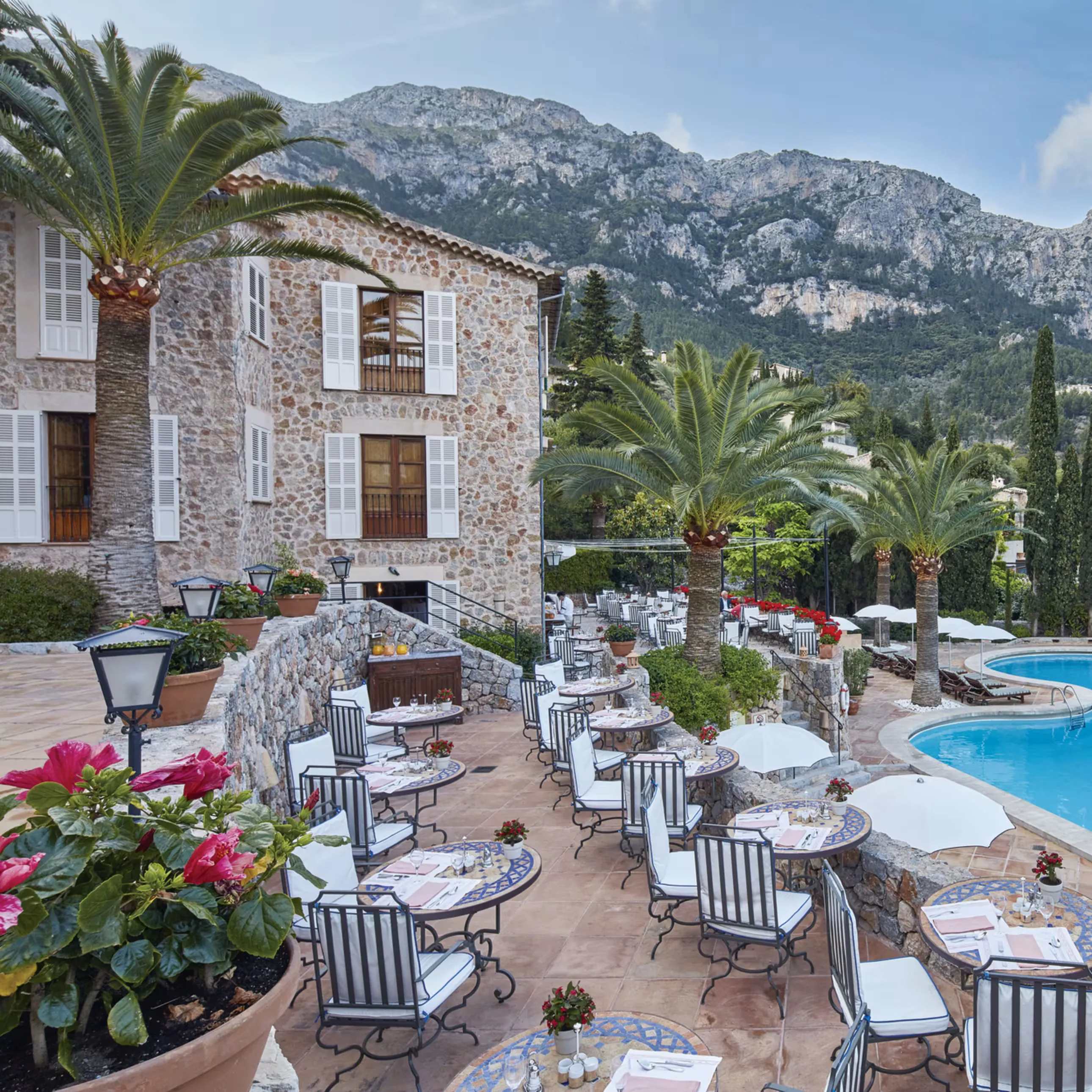 Restaurant terrace beside a stone manor house at Belmond La Residencia, with palms, the outdoor pool and the Tramuntana mountains behind