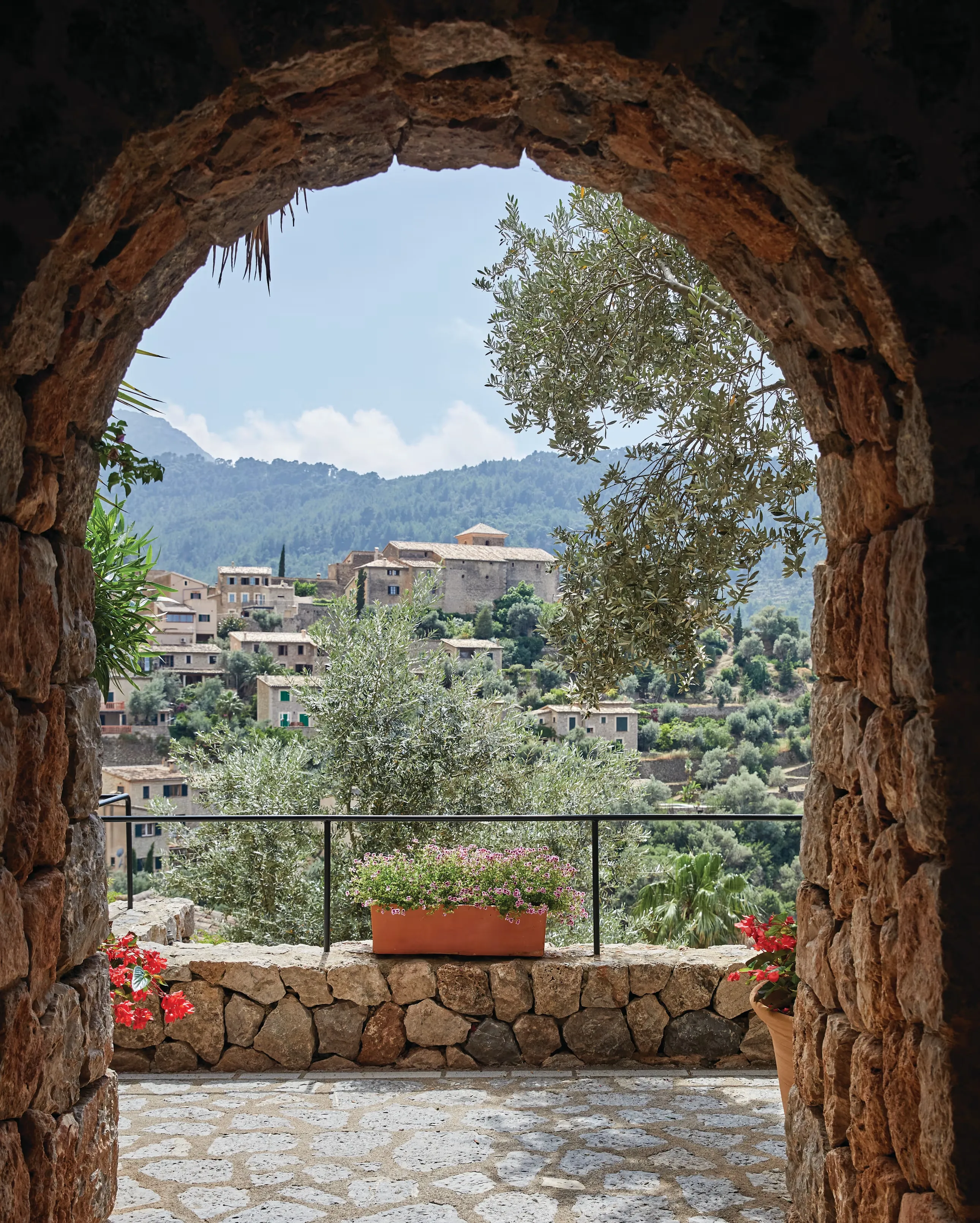 Deià seen through a rough stone arch: the village's honey-coloured houses climbing a hillside to a church tower, an olive tree and a terracotta trough of pink geraniums on the dry-stone wall in front