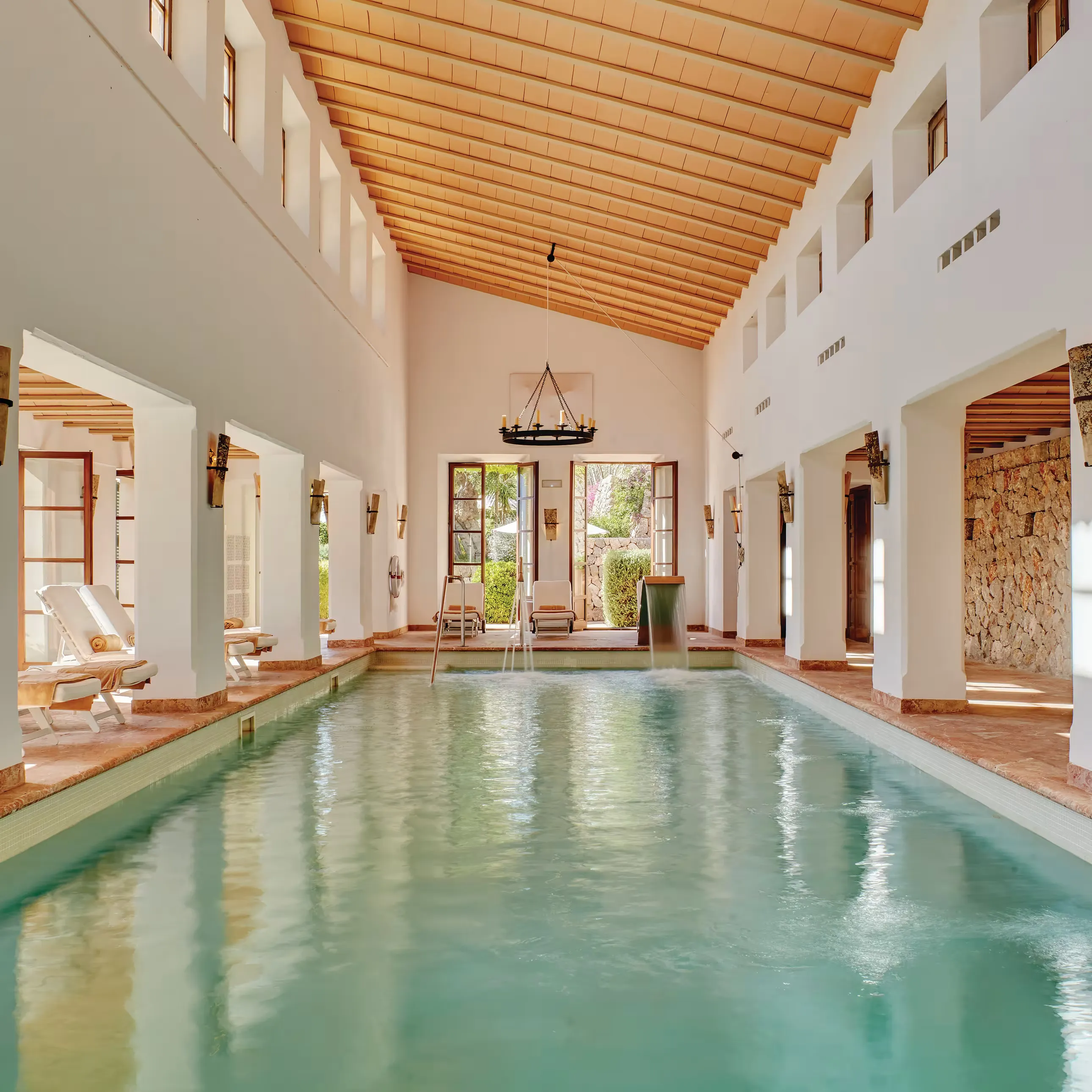 The indoor pool under a high pitched timber ceiling, with tall white walls, small deep-set windows, a wrought-iron candle chandelier and loungers in the arched bays along one side