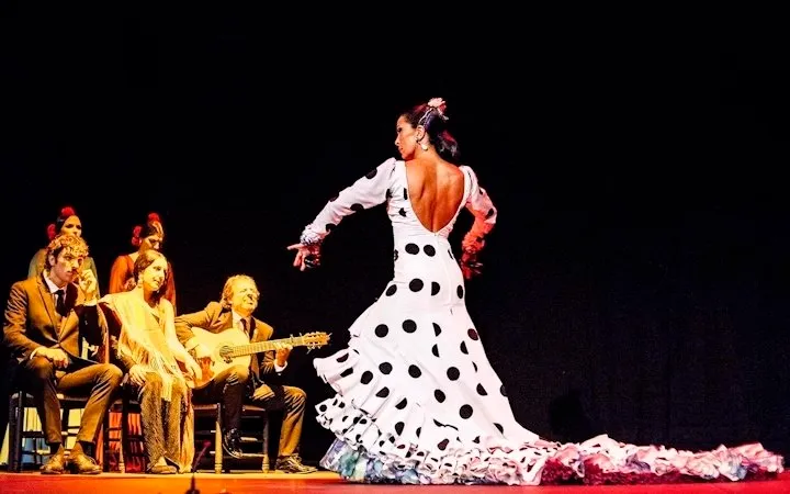 A flamenco dancer in a white dress with black polka dots and a long ruffled train turning on a stage, a guitarist, a singer and two women in shawls seated behind her