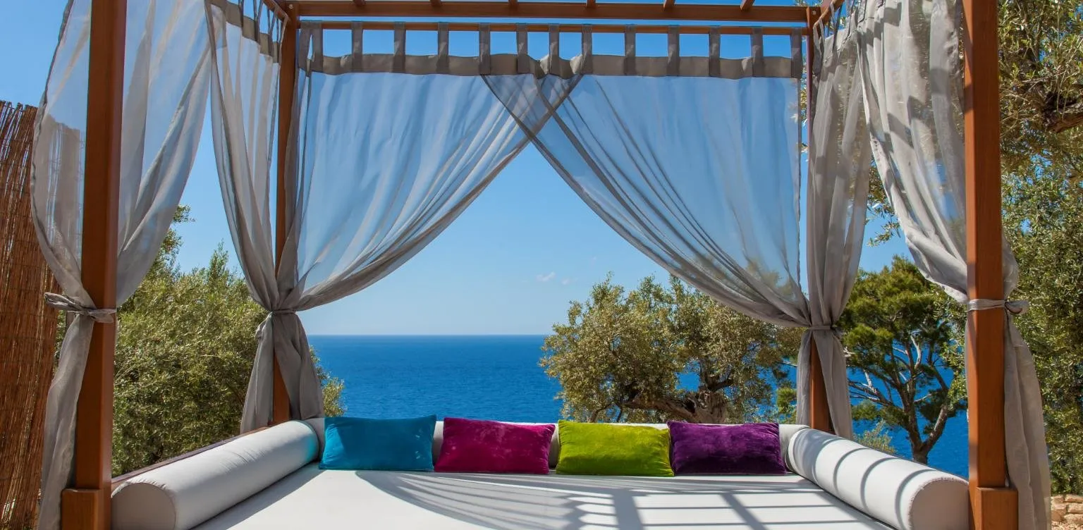 A daybed with white bolsters and four bright cushions under a wooden canopy hung with sheer grey curtains, olive trees and a deep blue sea beyond