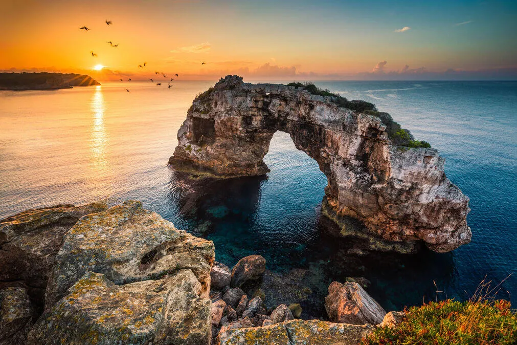 Es Pontàs, a natural stone arch in the sea, with birds over the water in the low sun
