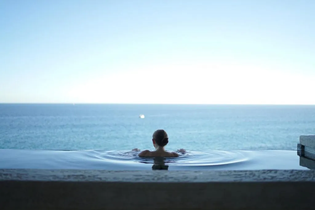 A woman seen from behind in an infinity pool, her arms on the rippling water, looking out over a calm blue sea to a pale horizon