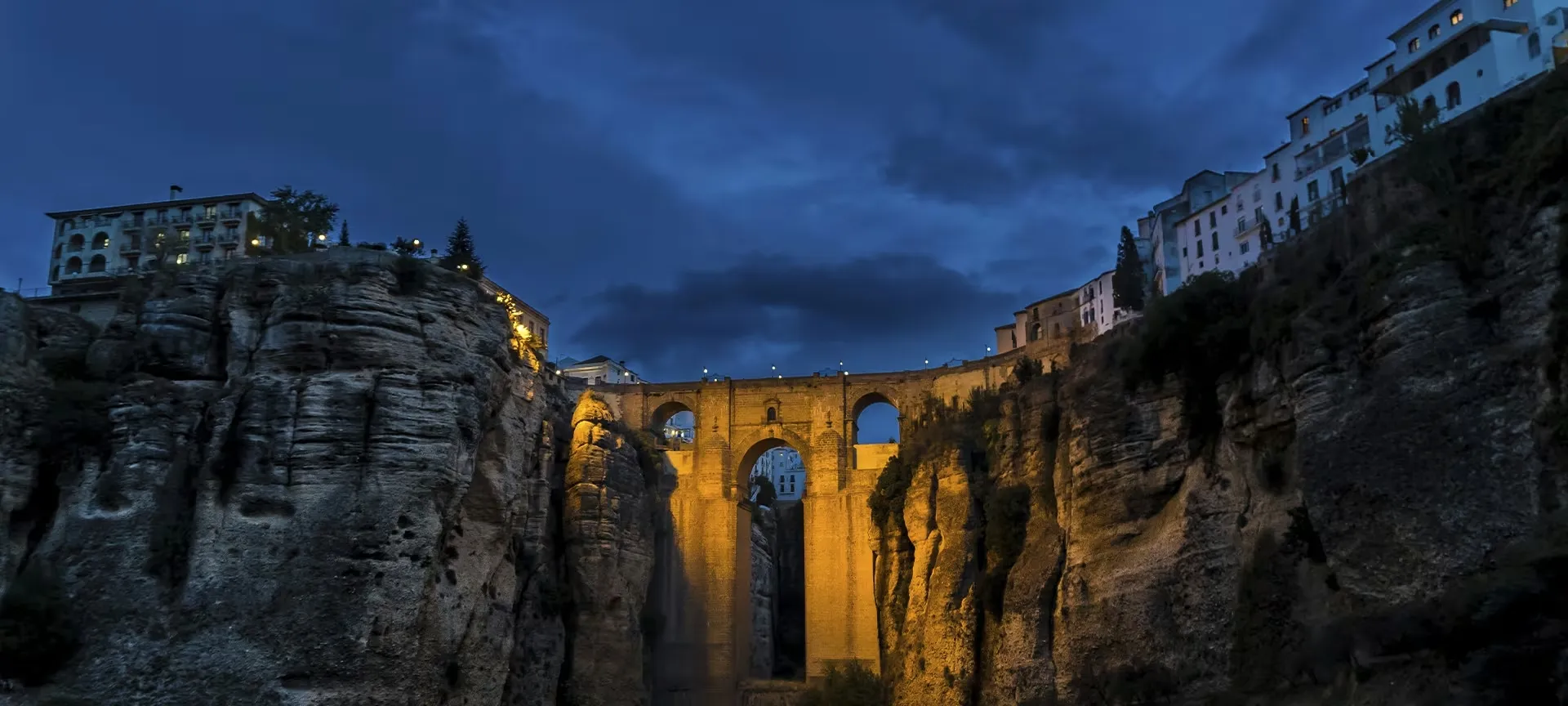 Ronda's Puente Nuevo lit gold at dusk, seen from the floor of the gorge between sheer rock walls, a large building on the cliff to the left and white houses along the clifftop to the right, under a deep blue sky