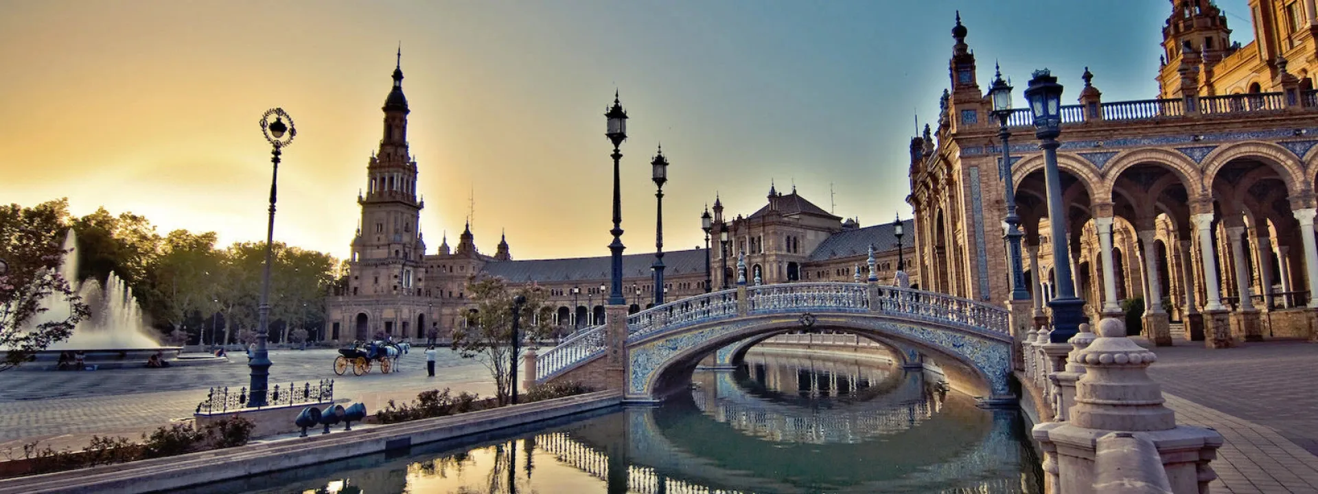 The Plaza de España in Seville at sunset: a tiled bridge arching over the canal, iron lamp posts, a tall tower and the curved arcades of the building, with a fountain and a horse-drawn carriage on the left