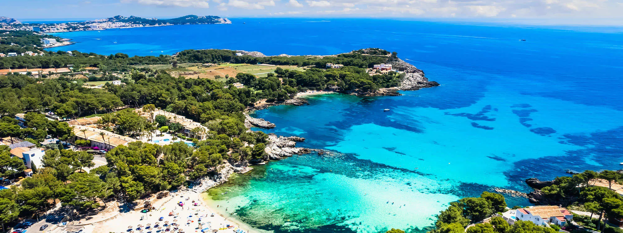 A cove on Mallorca from the air: turquoise shallows over sand and rock below pine woods and low houses, a beach of parasols in the foreground and a headland and town across the bay
