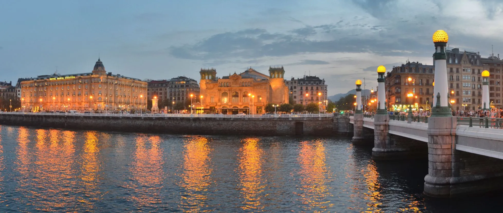 Hotel María Cristina, its name lit along the roof, and the Victoria Eugenia Theatre beside it glowing at dusk across the Urumea, their lights reflected in the river, with the globe lamps of a bridge at the right