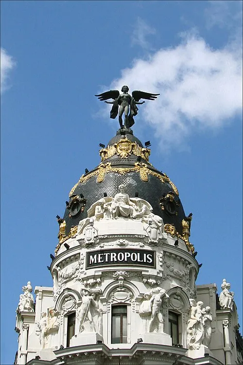 The dome of the Metropolis building in Madrid, crowned by a bronze winged figure, with the name METROPOLIS in gold letters beneath it, against a blue sky