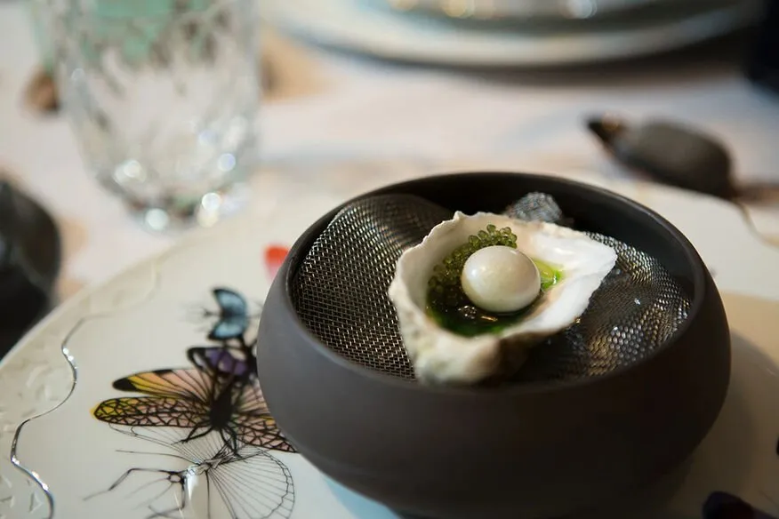 An oyster in its shell topped with a pearl-white sphere and green caviar-like beads, set on a mesh in a dark stone bowl on a plate painted with butterflies