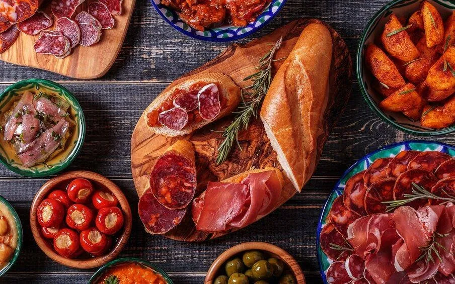 Spanish tapas from above on a dark wooden table: cured ham and chorizo on painted plates, bread on an olive-wood board, stuffed red peppers, green olives, anchovies and fried potatoes
