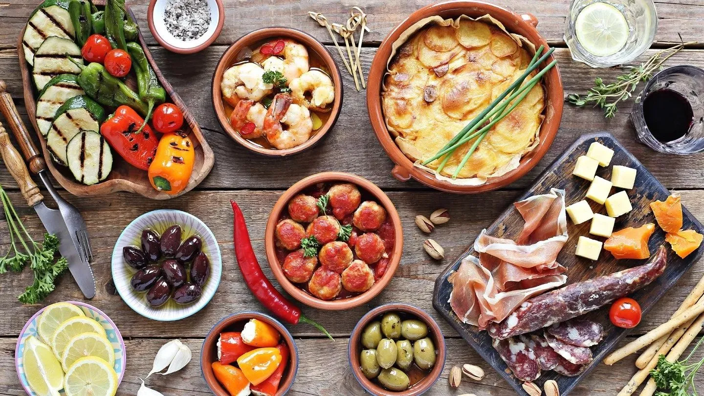 Spanish tapas from above on a weathered wooden table: prawns and meatballs in earthenware dishes, sliced potatoes baked in a clay pot, grilled vegetables, black and green olives, and a board of cured ham, sausage and cheese beside a glass of red wine