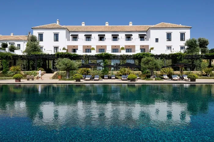 Finca Cortesín's white main building with its terracotta roof, seen across a long pool of clear blue water, with loungers and parasols along the terrace