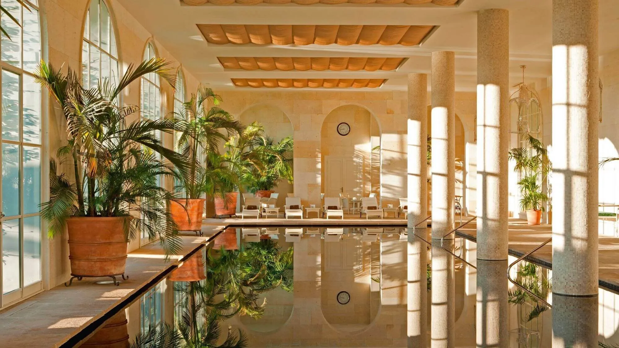A long indoor pool under a coffered ceiling, its still water reflecting tall arched windows, palms in terracotta pots, white loungers at the far end and a row of slender mosaic columns striped with sunlight