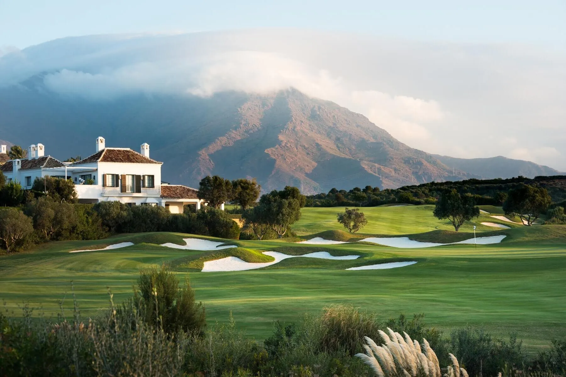 A white two-storey house with a terracotta roof, tall chimneys and green shutters among trees at the left, above a golf fairway set with white bunkers and a flag on the green, rough grass and pampas plumes in the foreground, and a reddish mountain half hidden in cloud behind