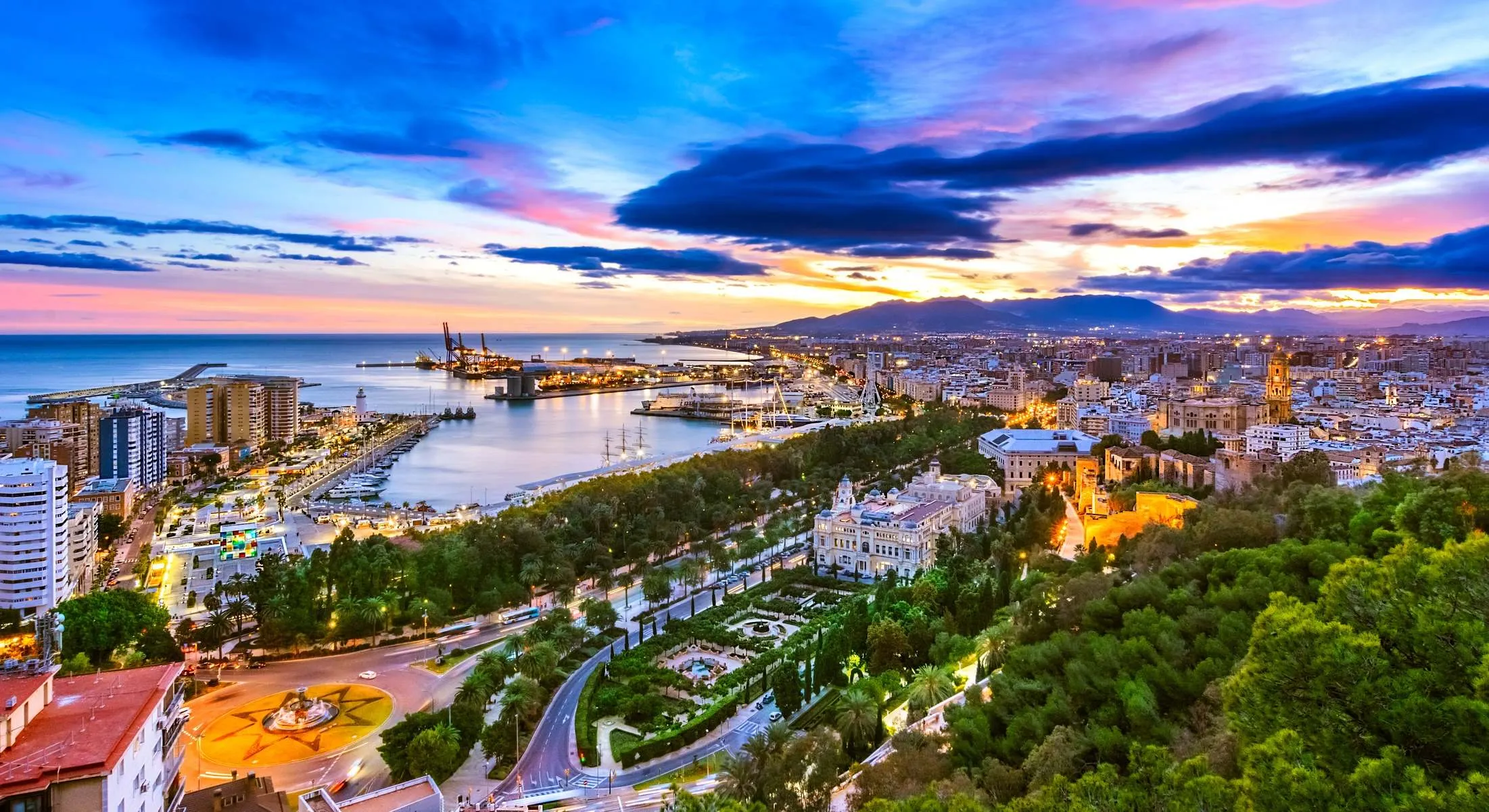 Málaga from the hill above it at dusk: gardens and lit streets below, and the port and the city spreading along the bay under a purple sky