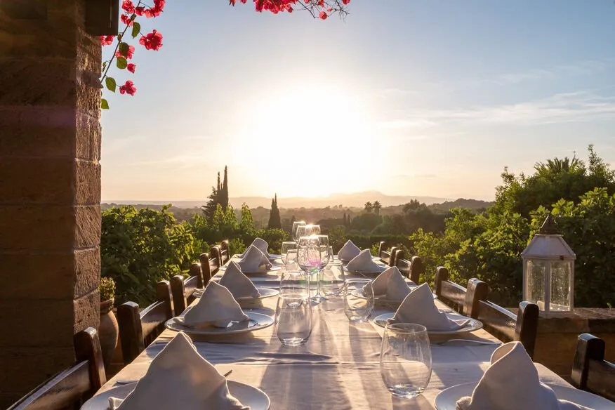 A long table laid with white linen, glasses and folded napkins on a terrace at sunset, bougainvillea overhead and gardens and hills beyond