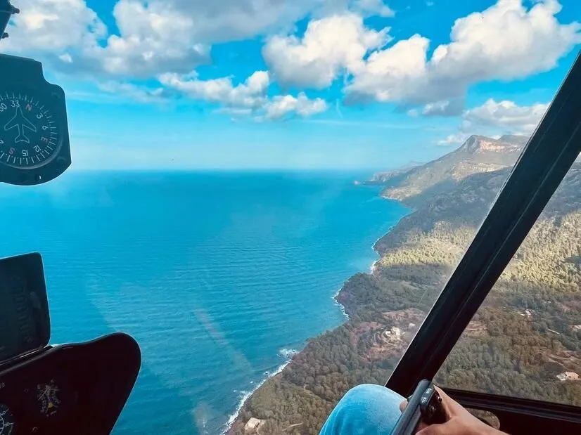 Mallorca's north-west coast seen from a helicopter cabin: wooded slopes falling to a deep blue sea and a headland in the distance, with a compass dial in the corner of the window