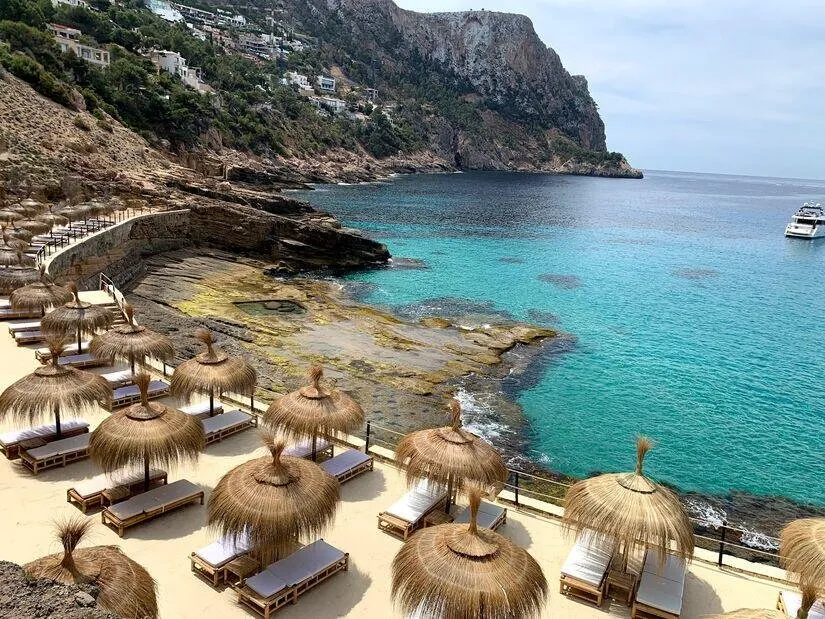 Thatched parasols and sunbeds on a terrace above the rocks of a cove, clear turquoise water below a steep wooded headland and a motor yacht at anchor