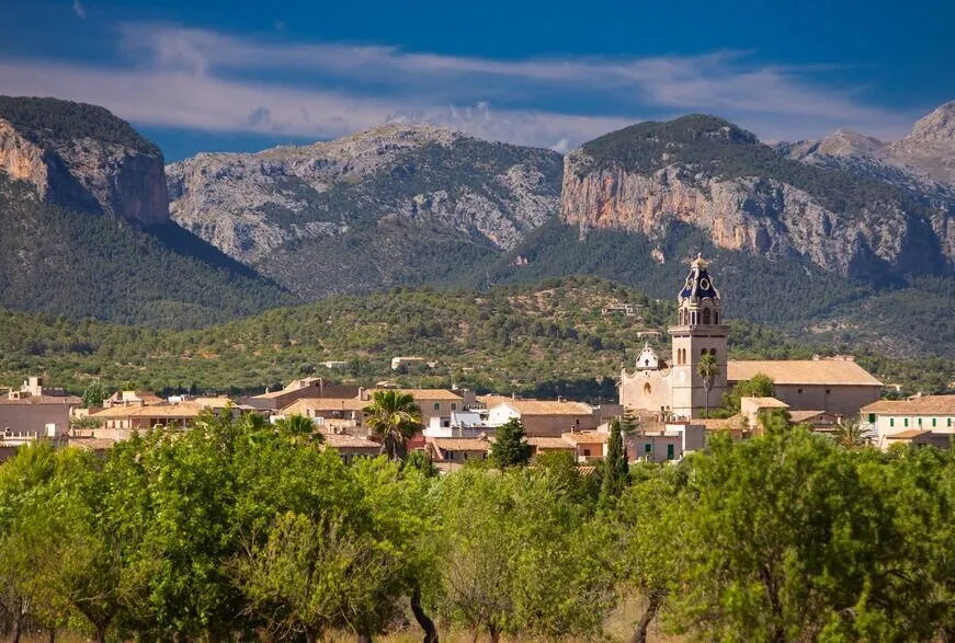 Santa Maria del Camí's church tower with its tiled dome above the village roofs and green trees, under the grey limestone cliffs of the mountains