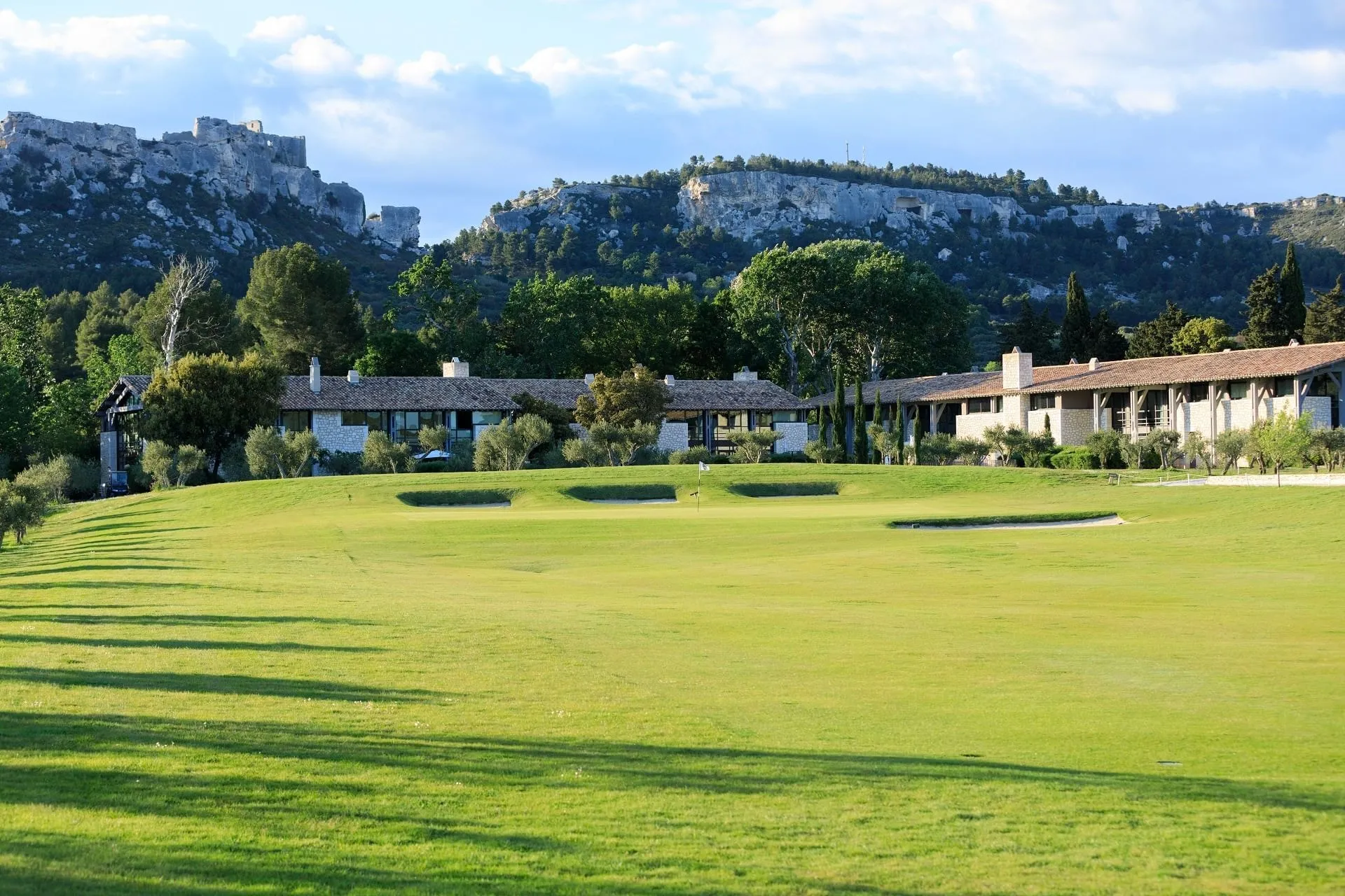 The estate's guest houses seen across a fairway: a long row of stone-and-render houses with terracotta-tiled roofs, square chimneys and tall glazed bays, olive trees and cypresses along their front, a green with a white flag and pale bunkers in the middle dist