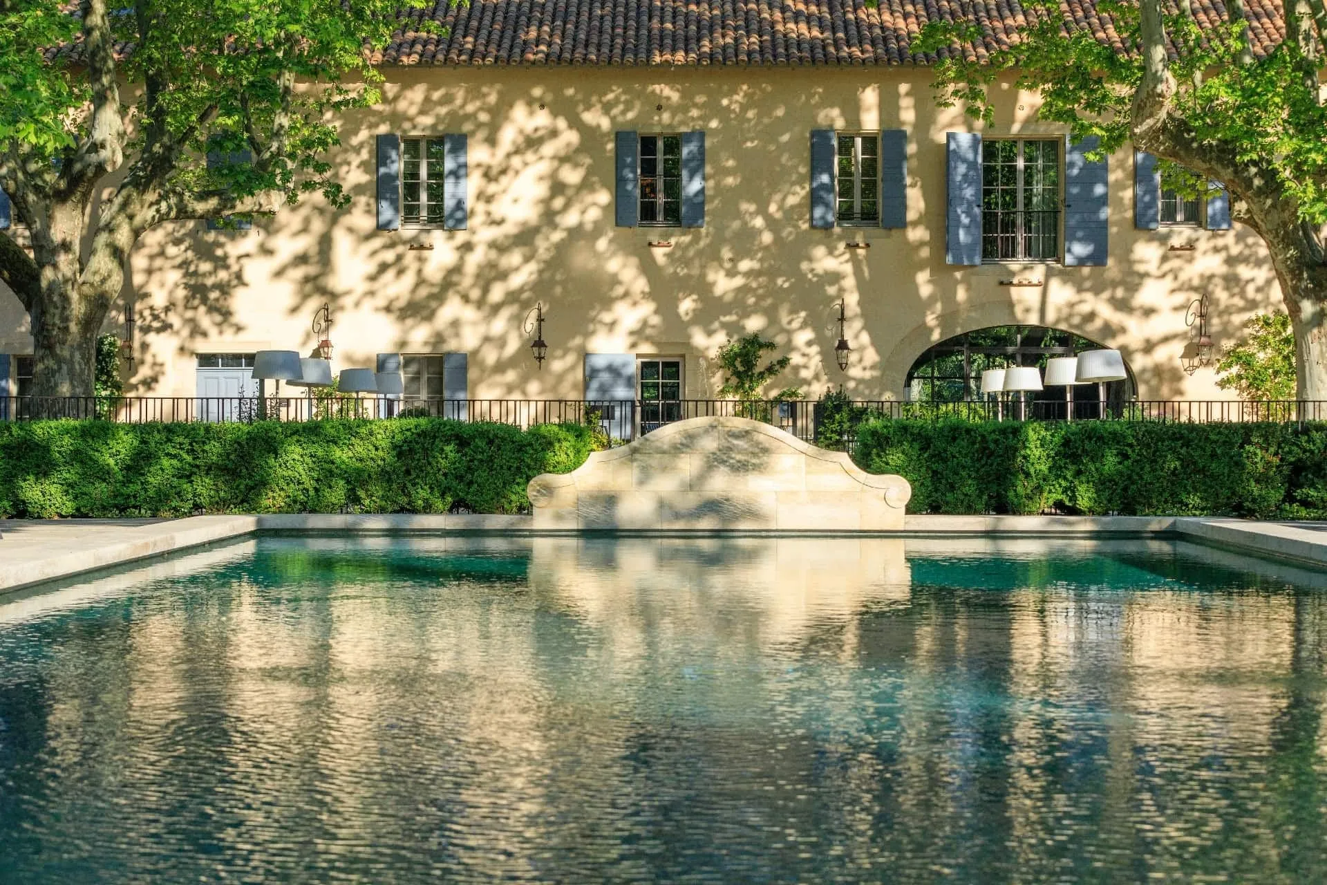 The main house's façade across the pool: a long ochre-rendered two-storey building with a terracotta roof, blue-grey shutters, iron lanterns and an arched doorway, in the dappled shade of plane trees; a clipped box hedge and an iron railing along the pool's fa