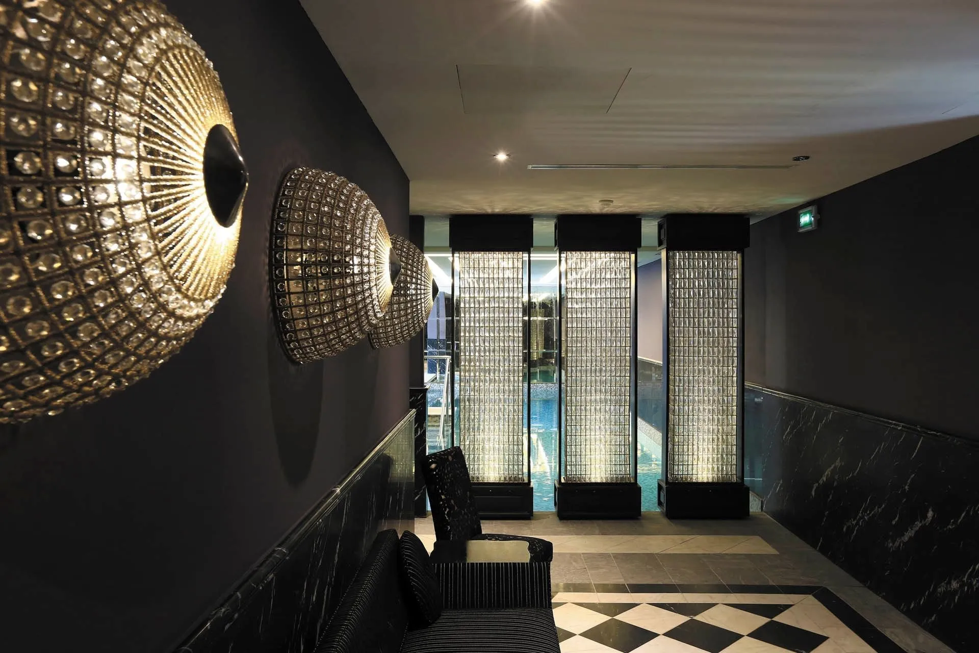 The spa's corridor to the indoor pool: dark grey walls with three domed crystal-beaded sconces lit warm, a black-and-white marble dado, a chequered marble floor, a black striped chaise and a black armchair; at the end, three lit glass-brick pillars frame the t