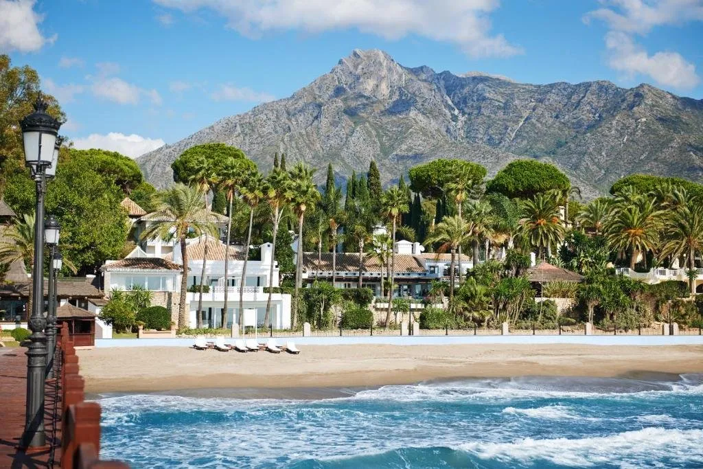 A sandy beach and white low-rise buildings among palms and pines, seen from a pier over the waves, with a bare rocky mountain rising behind under a blue sky