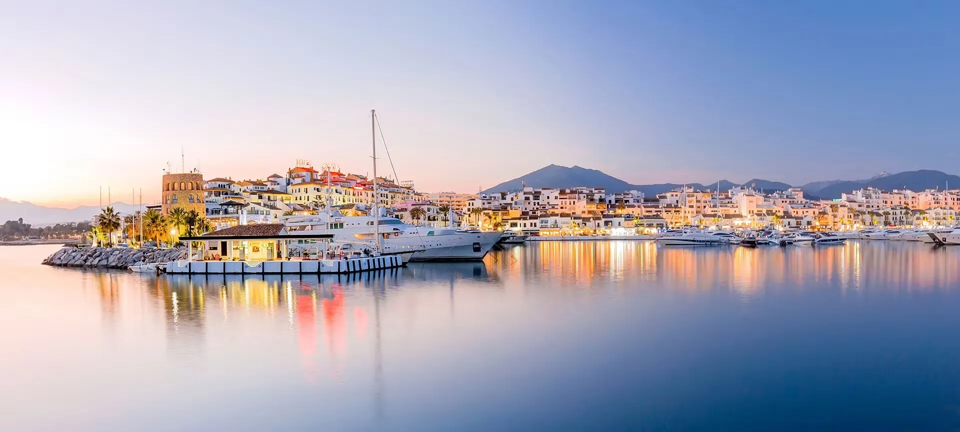 A marina at dusk: yachts moored along a lit waterfront of white buildings and a stone tower, their lights reflected in still water, mountains behind under a pale violet sky