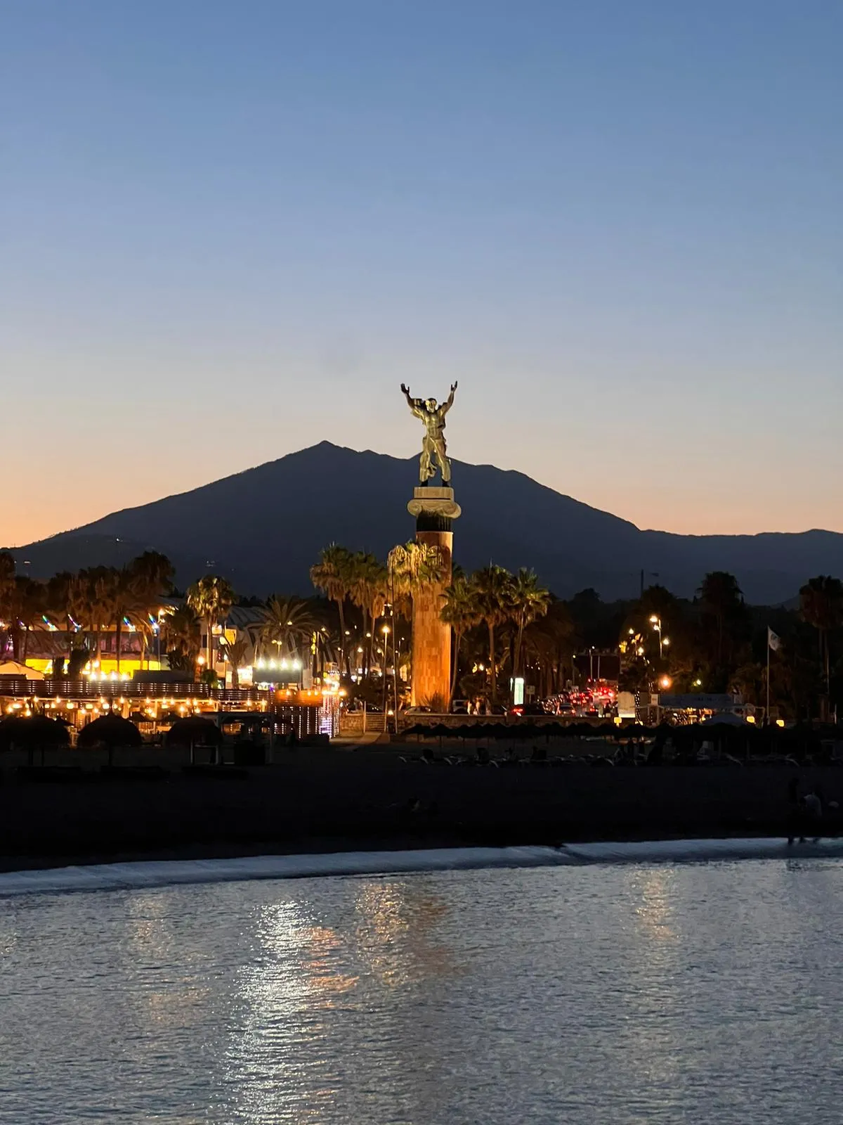 A bronze figure with raised arms on a tall column, lit gold at dusk above a promenade of palms and lights, a mountain dark against the evening sky behind it and the calm sea in front