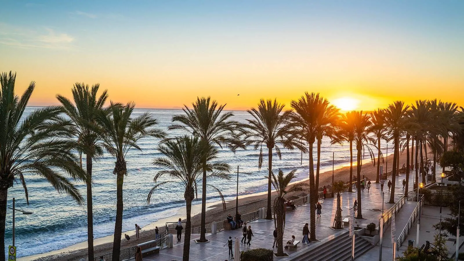 A promenade of tall palms above a sandy beach at sunset, people strolling along it, the sun low over the sea on the right and the sky turning gold
