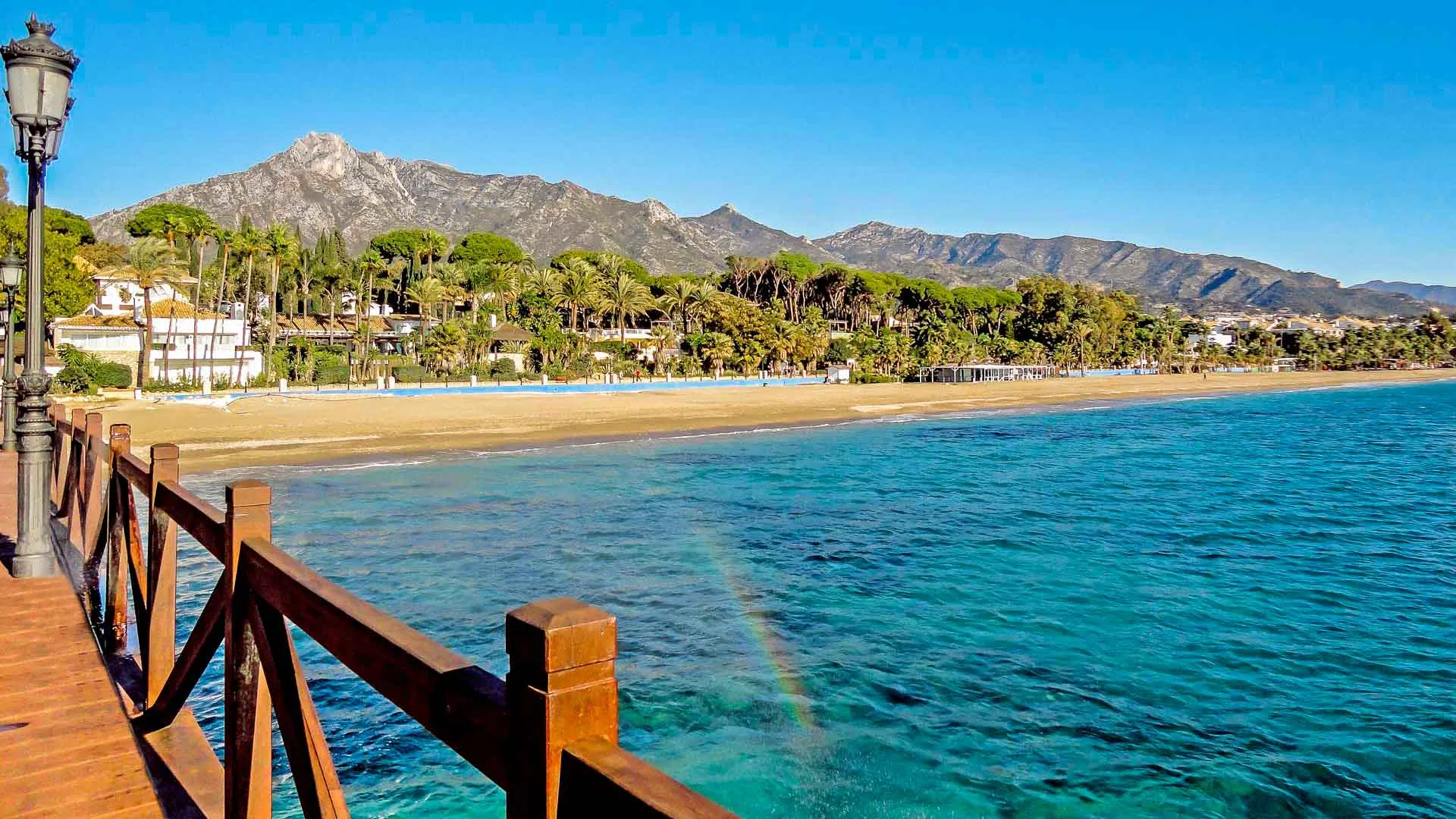 The coast at Marbella from a wooden pier: clear turquoise water, a long sandy beach lined with palms and white villas, and the rocky peaks of Sierra Blanca behind under a cloudless sky