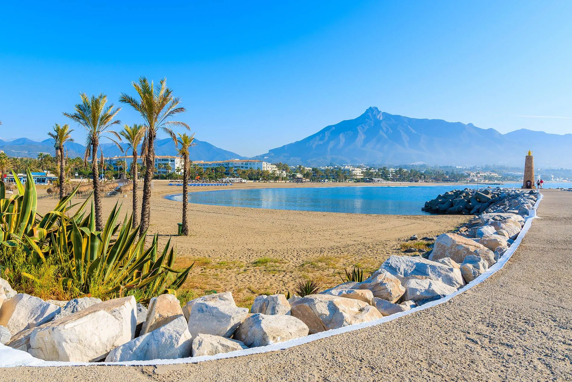 A beach of pale sand with palms and spiky plants, a curved breakwater of white rocks with a small lighthouse, calm blue water and a sharp grey peak behind